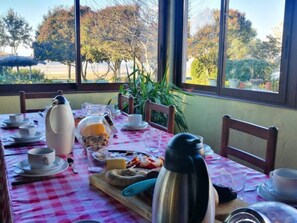 A dining table set with plates, cups, and a kettle, with a view of trees and a building in the background.