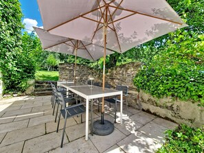 Eine Terrasse im Freien mit Tisch und Stühlen unter einem großen Sonnenschirm, umgeben von Pflanzen und Steinmauern.