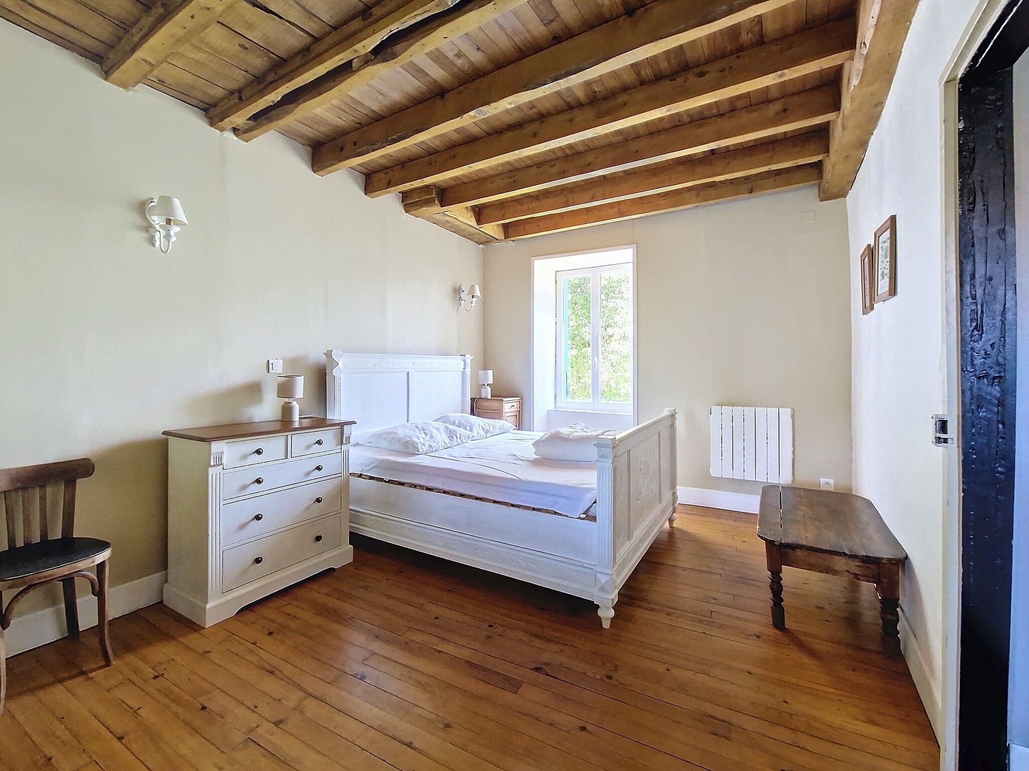 Un dormitorio con techo de madera, cama con ropa de cama blanca, cómoda, mesita de noche y ventana con vista a zonas verdes.