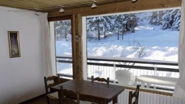 A cozy room with a wooden table and chairs, a radiator, and a view of a snowy landscape through large windows.
