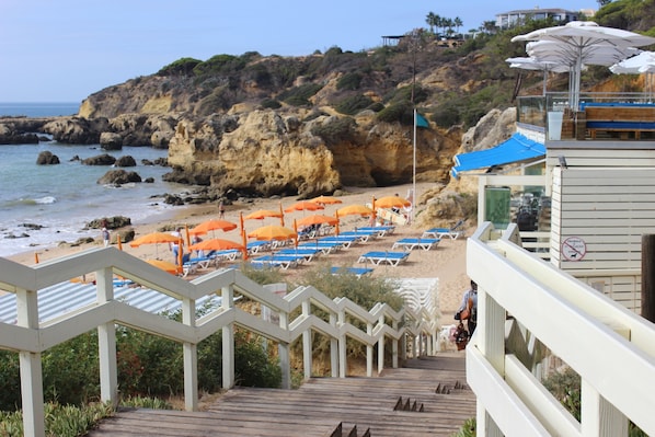 A beach with wooden stairs leading down to the shore, featuring orange umbrellas and blue lounge chairs.