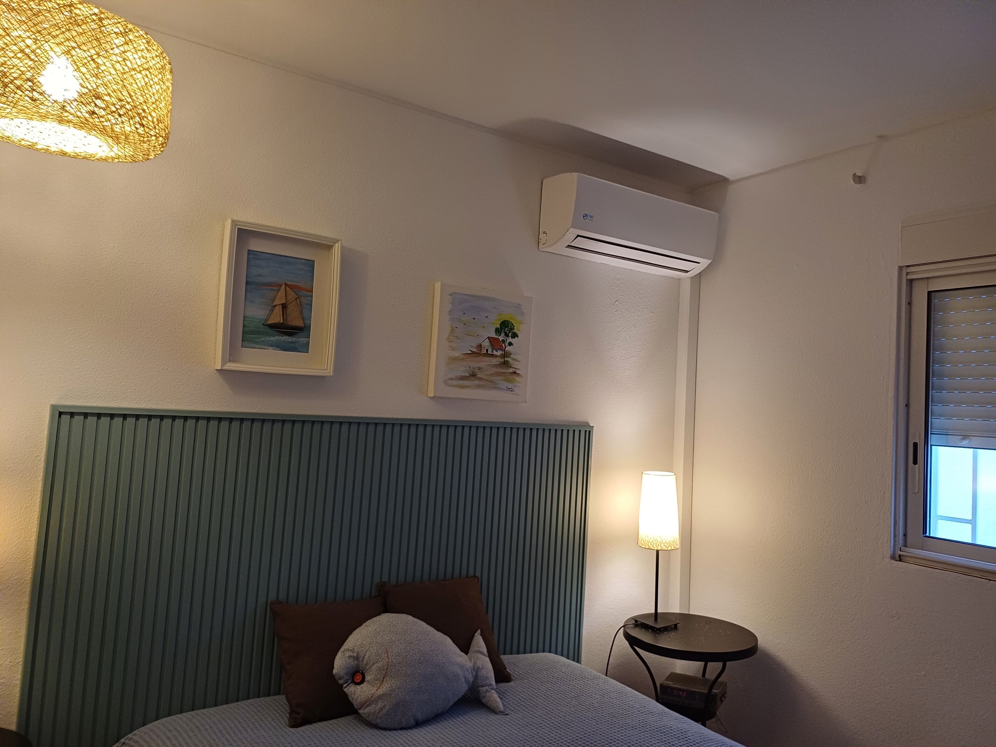 A hotel room with a bed, a wall-mounted air conditioner, two framed pictures, a lamp, and a window with blinds.