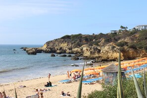 A beach with sun loungers and umbrellas, a rocky cliff, and a building in the background.