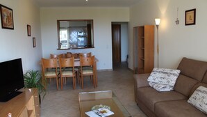 Una sala de estar con un sofá, una mesa de centro de vidrio, un televisor, una zona de comedor con sillas y una mesa, y una cocina visible a través de una ventana.