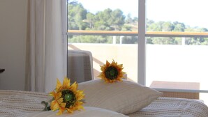 Habitación de hotel con cama, dos almohadas con girasoles, ventana con vista a árboles y barandilla de balcón.