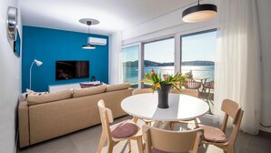 A modern living room with a large TV, a sofa, a dining table with chairs, and a view of the sea through large windows.