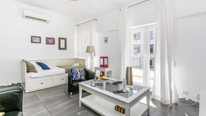 Une pièce moderne et bien éclairée, avec un canapé blanc, une table basse en verre et une vue sur l’extérieur grâce à de grandes fenêtres.