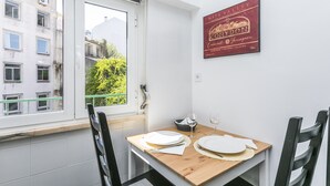 Un coin repas avec une table dressée pour deux personnes, une fenêtre donnant sur des bâtiments et de la verdure, et un panneau encadré accroché au mur.
