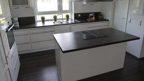 Eine moderne Küche mit weißen Schränken, einer schwarzen Arbeitsplatte und einer großen Kochinsel. Über der Spüle befindet sich eine Mikrowelle, links ein Herd und ein Fenster, das für natürliches Licht sorgt.