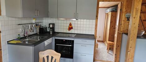 A compact kitchen with grey cabinets, a wooden ceiling, a dining table with a white tablecloth, and a green chair.