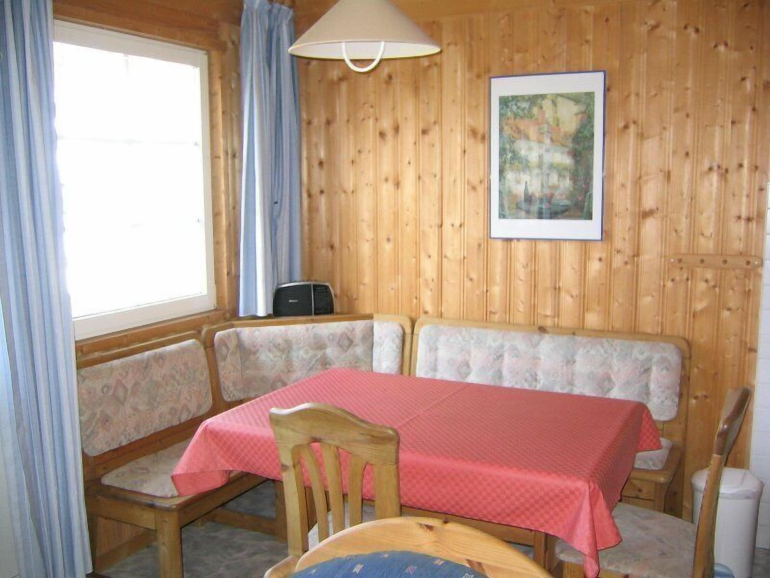 A wooden cabin with a dining table and chairs, a window with curtains, and a framed picture on the wall.