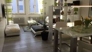 A modern living room with a sofa, a glass table, and a dining area with a chair and a lamp. There are potted plants and a radiator in the room.