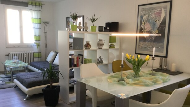 A modern living room with a glass dining table, white chairs, a bookshelf, and a radiator.