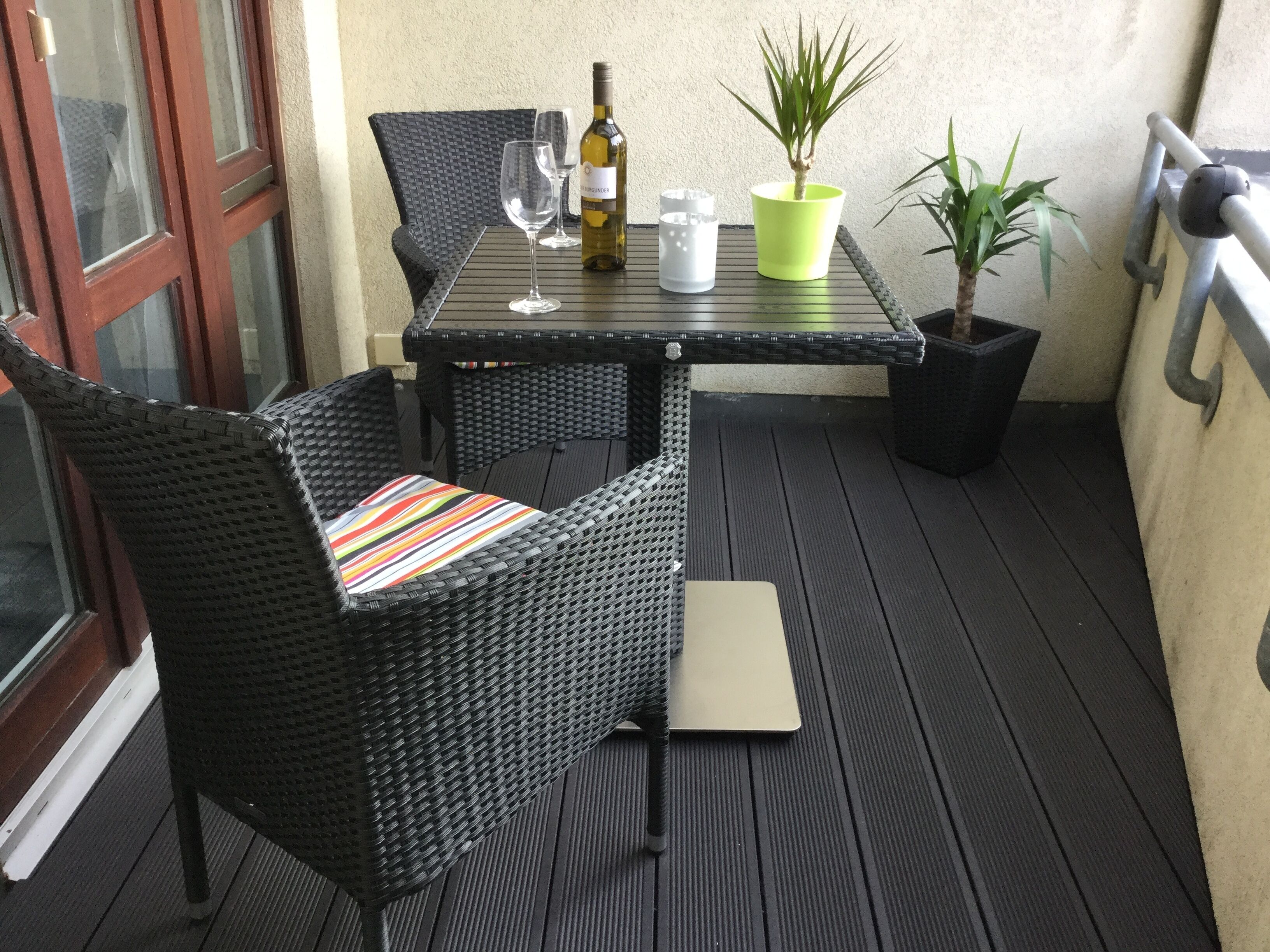 Eine kleine Terrasse im Freien mit Korbmöbeln, einem Tisch, einer Weinflasche, einem Glas und einem Blumentopf.