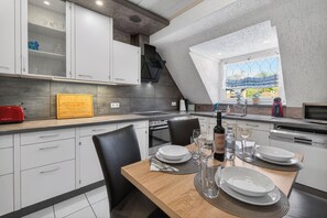 A modern kitchen with white cabinets, a wooden dining table set with plates, glasses, and cutlery, and a view of greenery through a window.