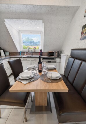 Un comedor con una mesa dispuesta para cuatro personas, una cocina al fondo y vistas a través de una ventana.
