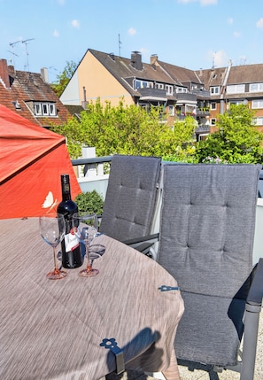 A table with a bottle of wine and two glasses, outdoor seating, and a red umbrella.