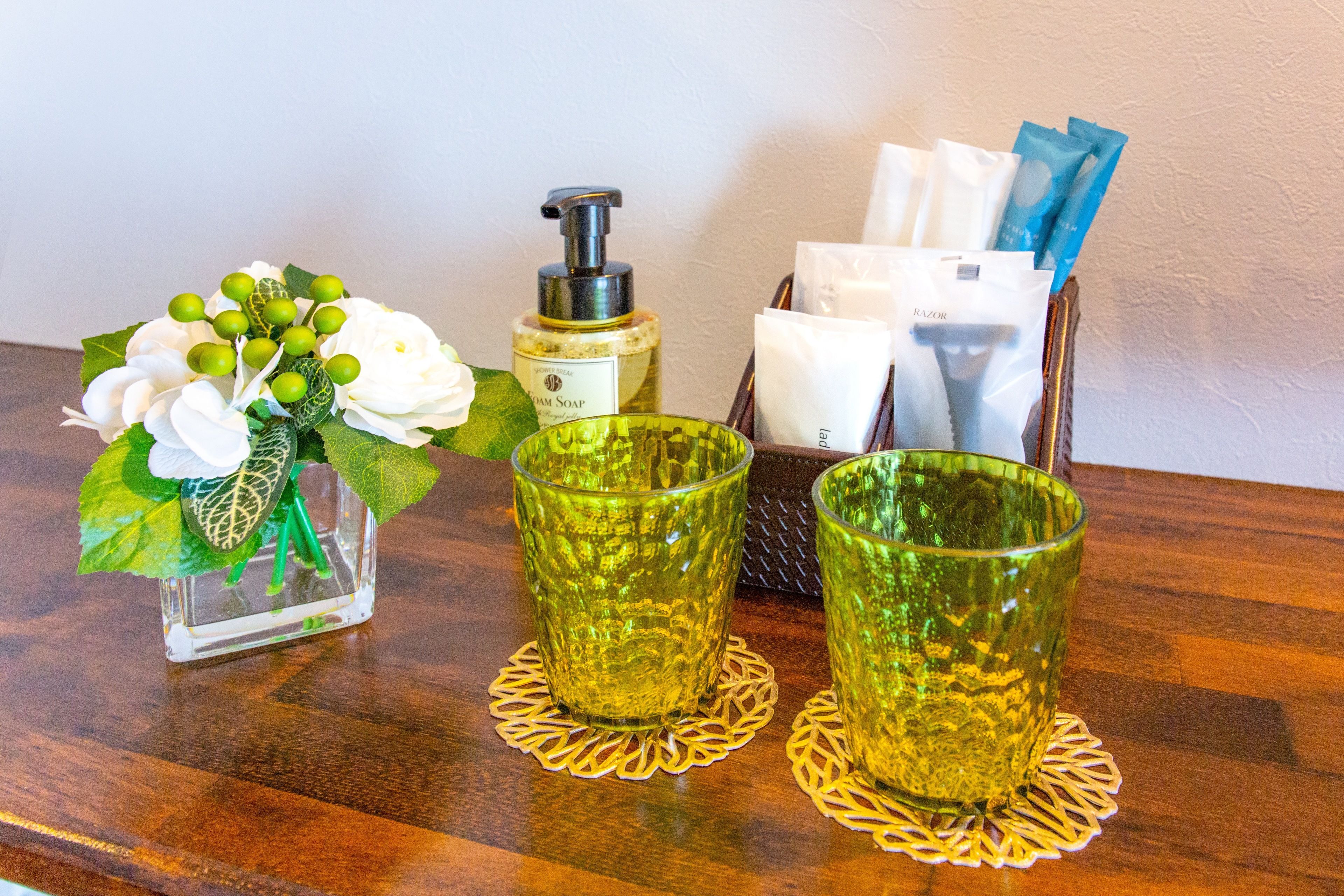 A wooden table with a clear glass vase containing white flowers and green leaves, two green glass cups with a textured pattern, a bottle of soap, and a tissue holder with tissues and a blue item.
