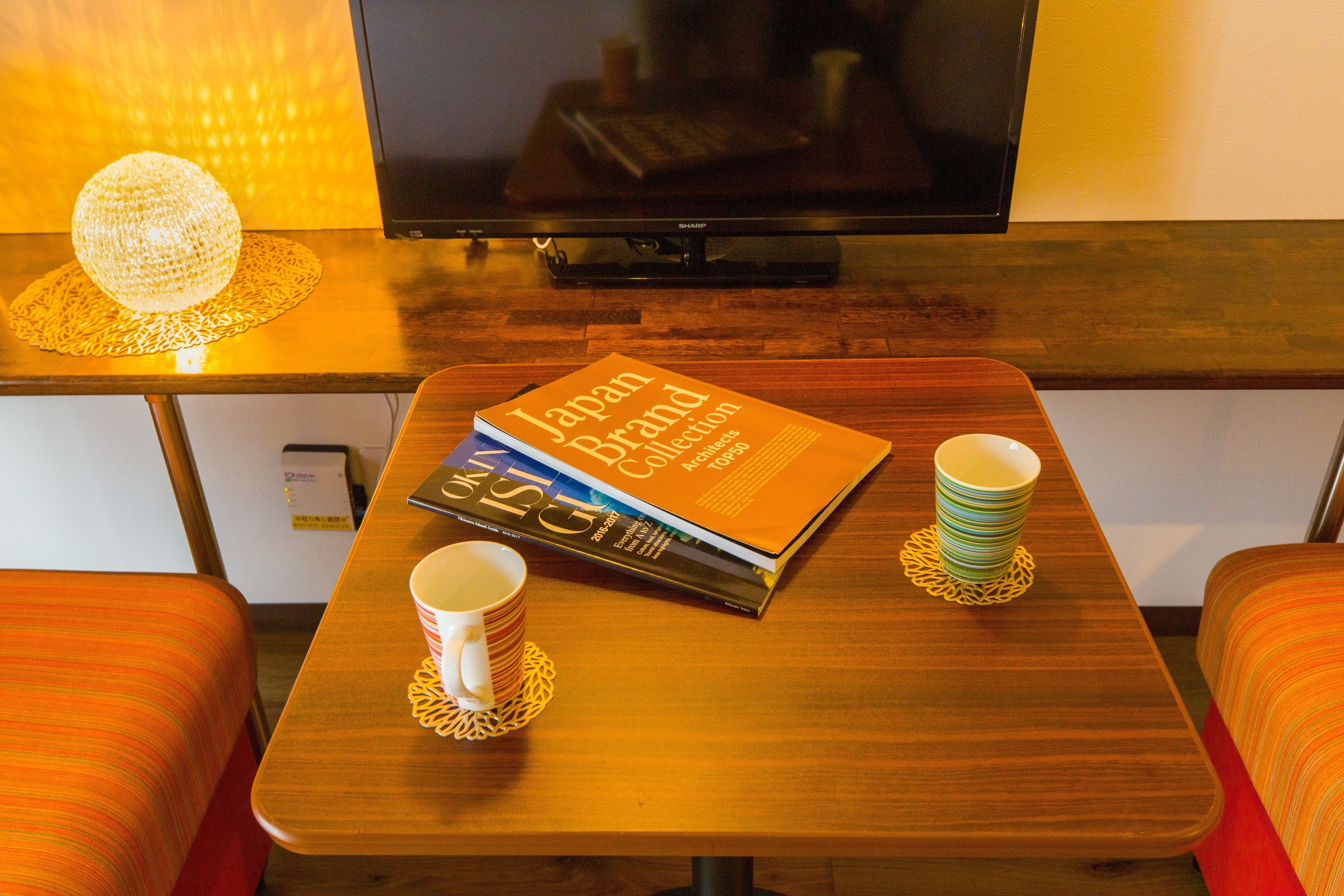Ein Hotelzimmer mit einem Holztisch, einem Buch mit dem Titel "Japan Brand Collection", zwei übereinander gestapelten Tassen und einem an der Wand montierten Fernseher.