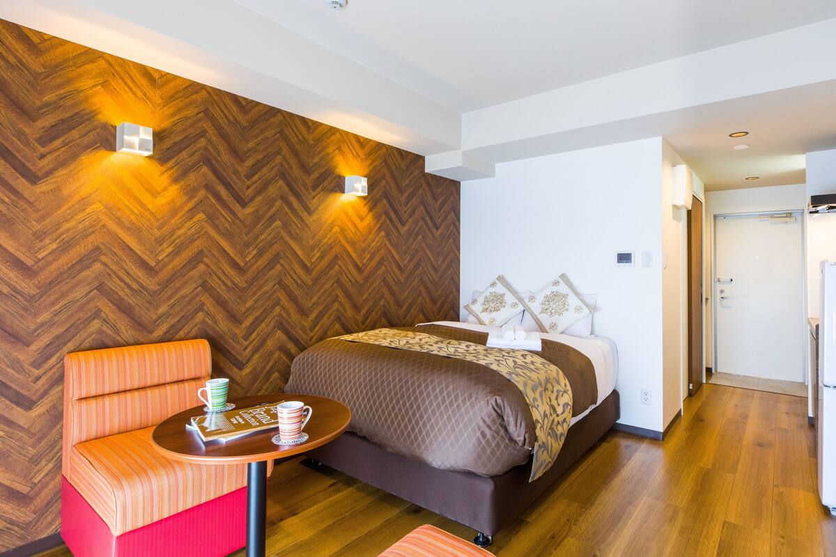 A hotel room with a wooden herringbone wall, a bed with a brown quilt, a small table with a cup and a book, and a red and orange chair.
