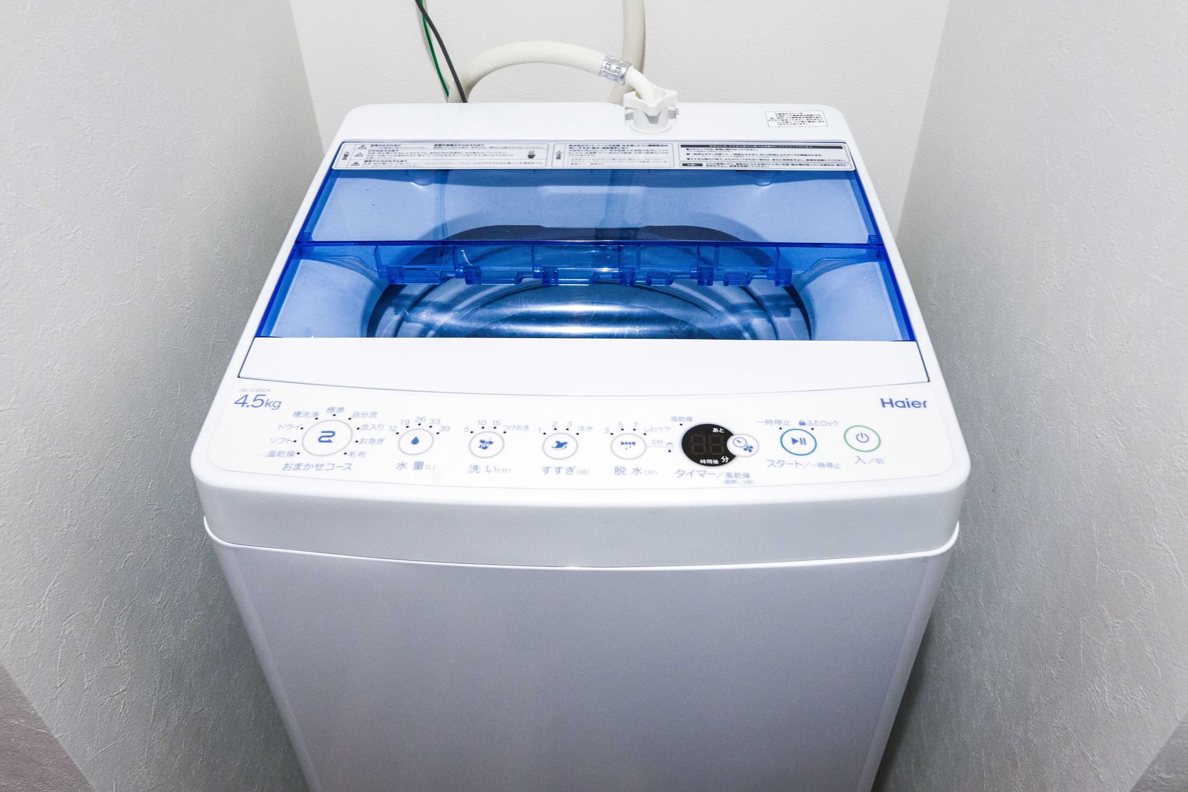 Vista frontal de una lavadora Haier blanca con puerta transparente, panel de control con varios botones y un cable conectado a la pared.