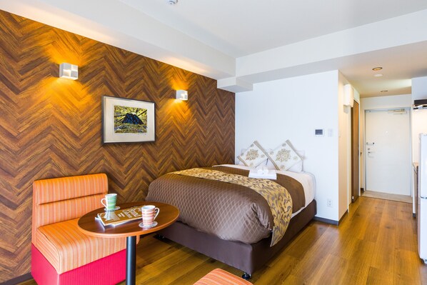 A modern hotel room with a wooden herringbone wall, a bed with a brown quilt, a small table with a cup, and a red and orange chair.