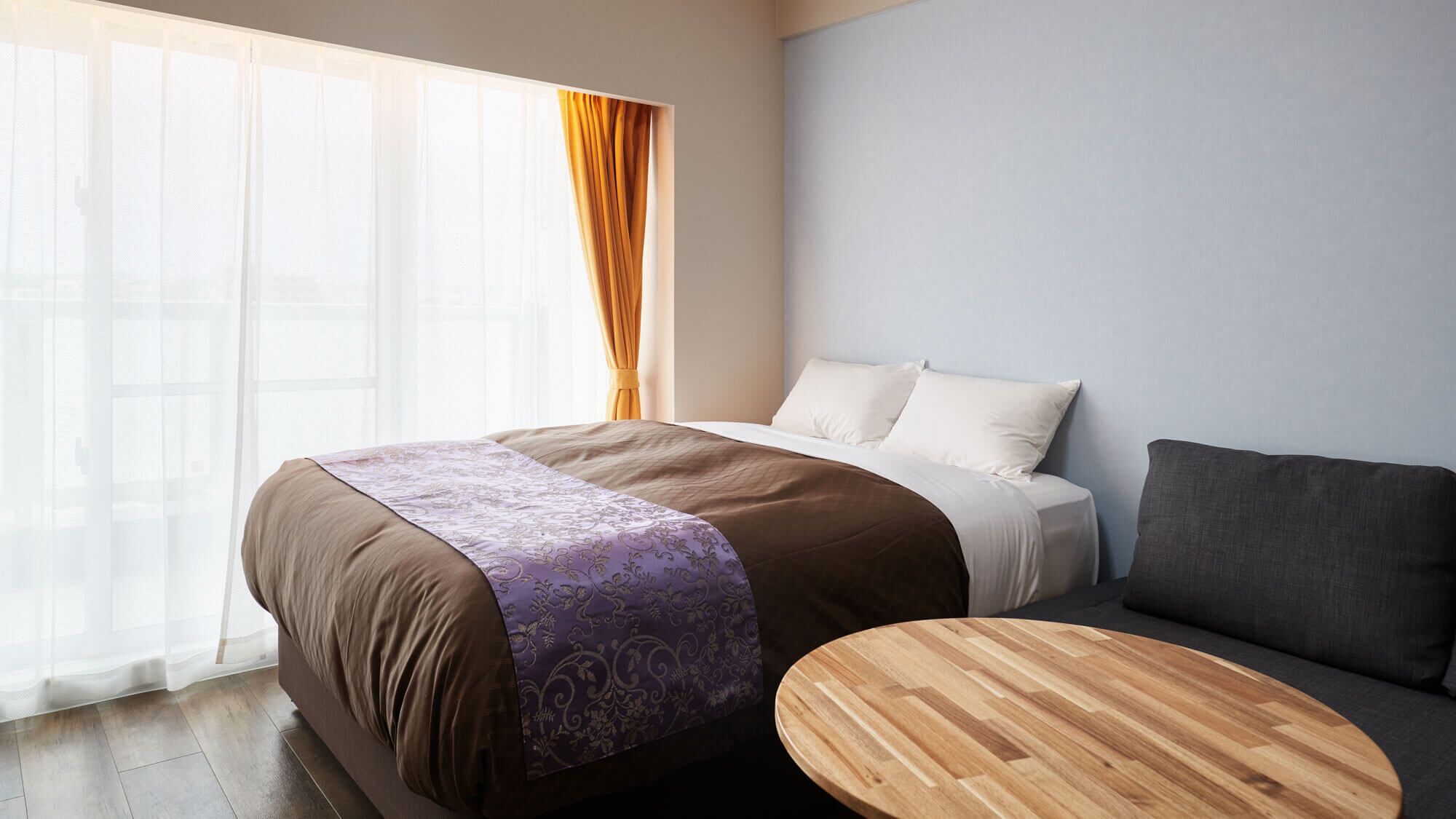 A hotel room with a bed, a sofa, a round wooden table, and a large window with sheer curtains.