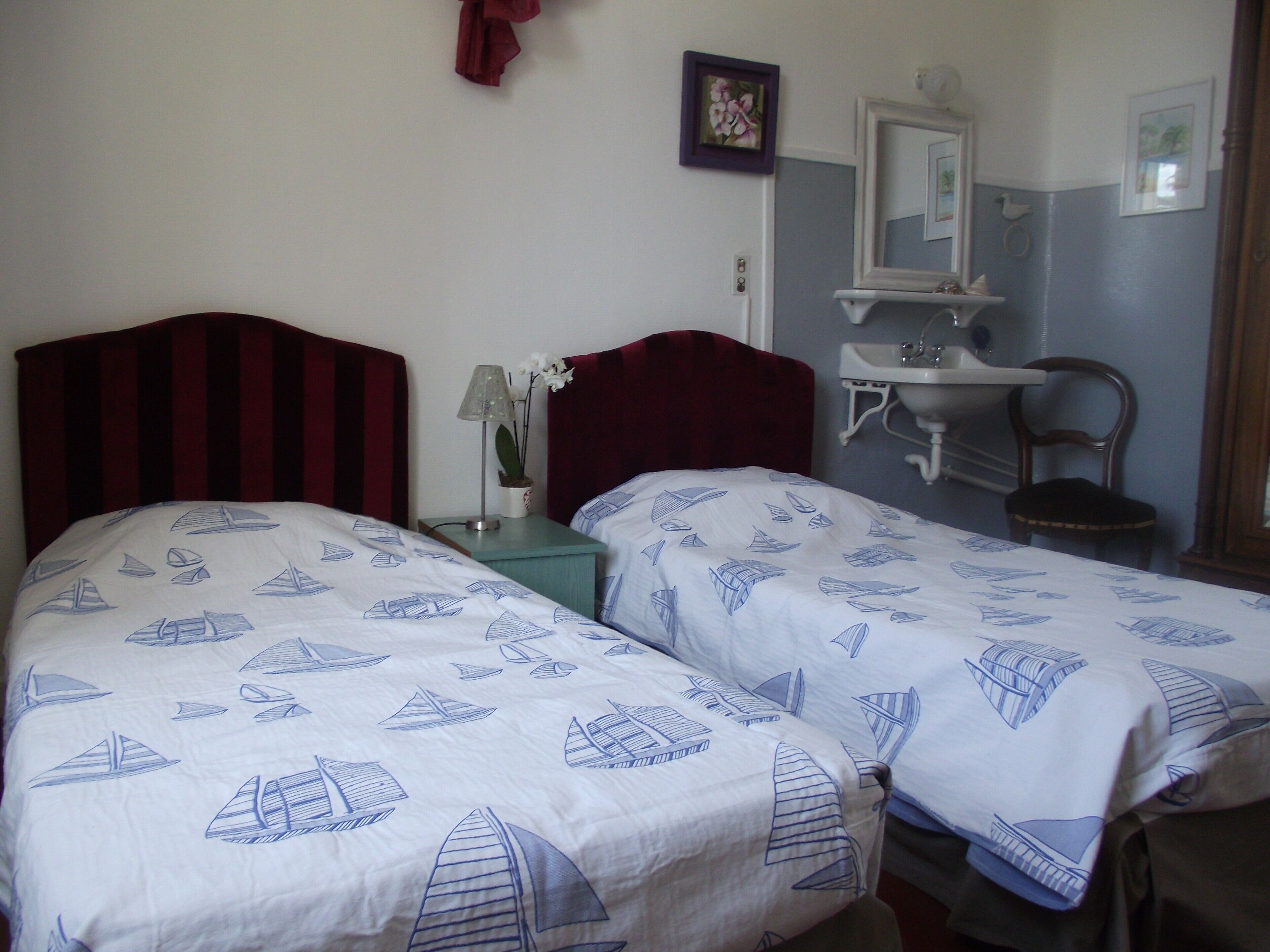 Habitación de hotel con dos camas, una mesita de noche, una lámpara, un florero con flores, un espejo, un lavamanos y una silla.