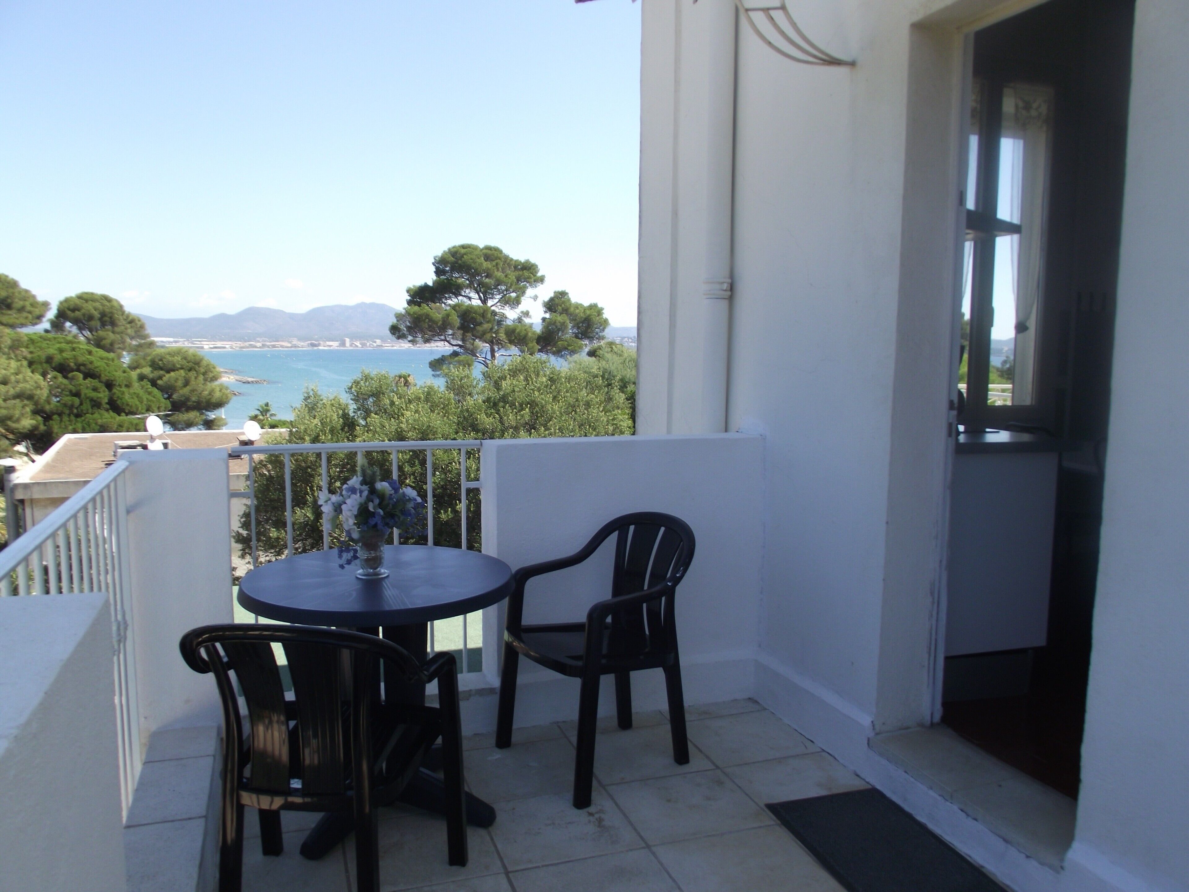 Un balcone con tavolo e due sedie, affaccia su un paesaggio costiero con alberi e vista sul mare.