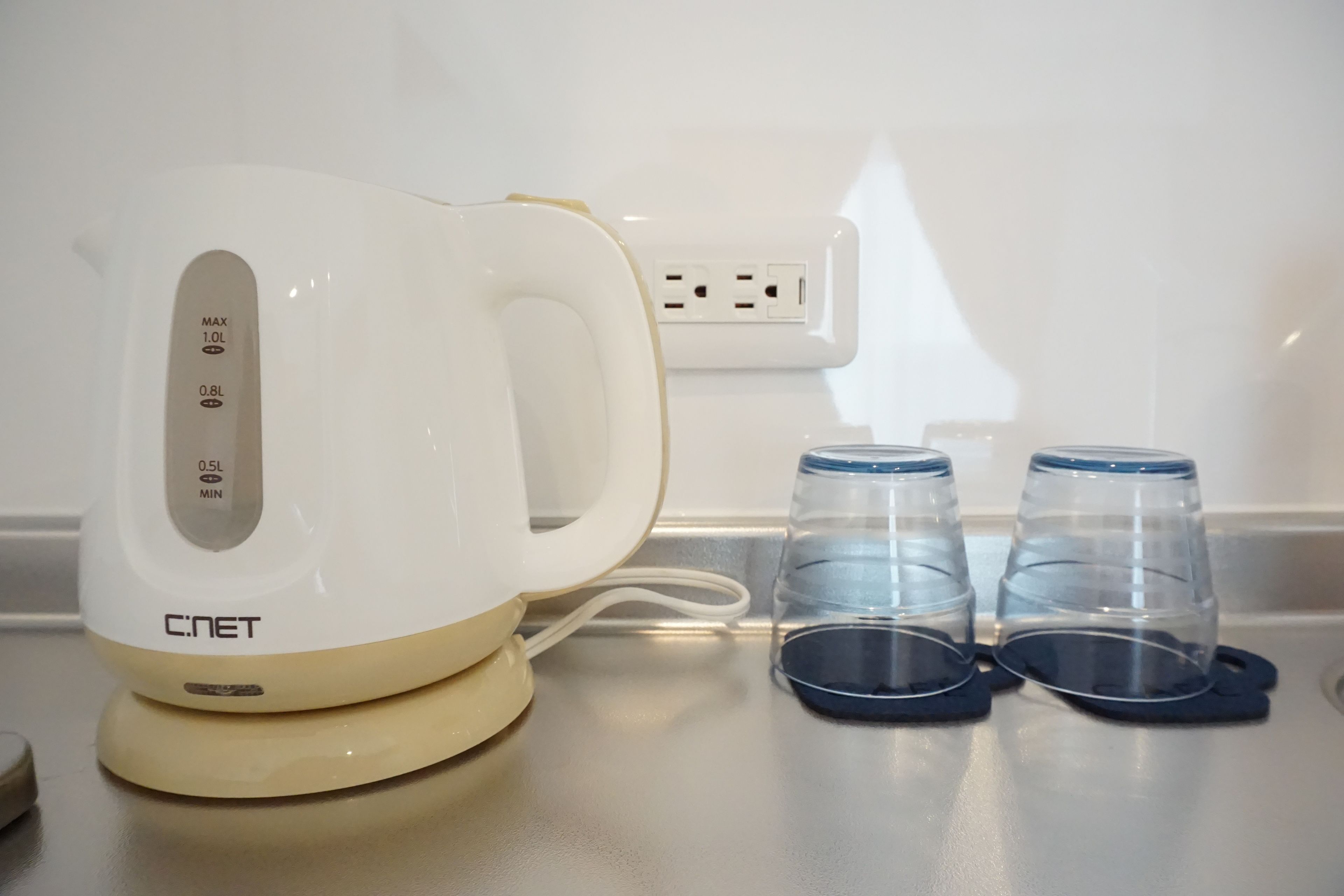 A white electric kettle with a digital display, two clear glasses with blue rims, and a power outlet in the background.