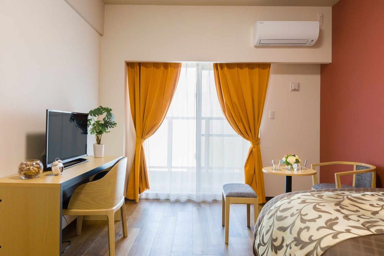 A hotel room with a bed, a desk with a TV, a chair, a small table with a vase, and a window with curtains.