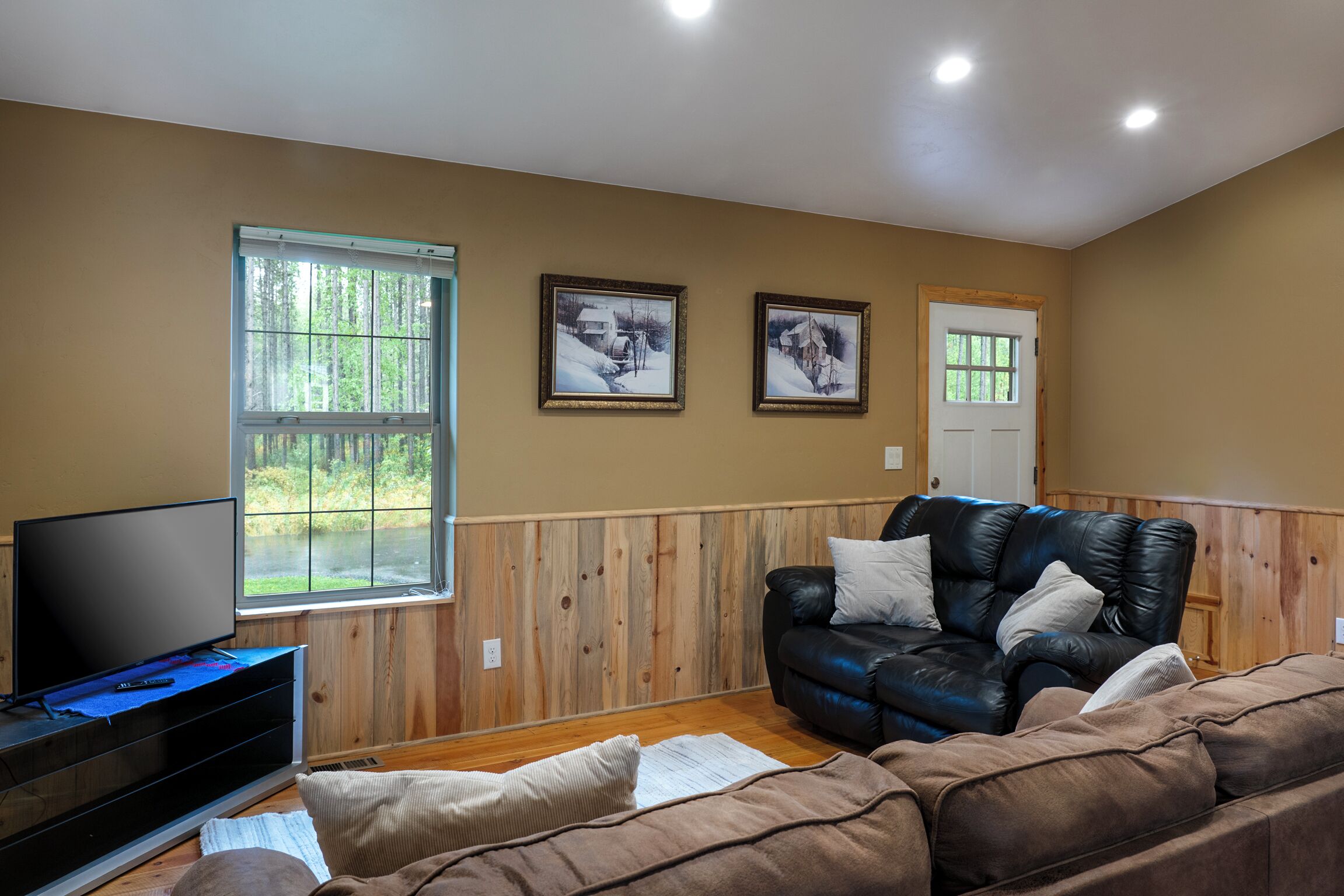 A living room with a television, wooden paneled walls, a black leather sofa, and two framed pictures on the wall.