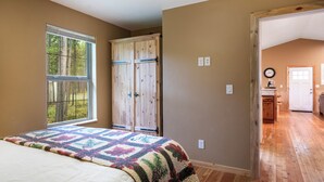 Ein Schlafzimmer mit einem großen Bett, einem Fenster mit Blick auf Bäume, einem hölzernen Kleiderschrank und einem Flur, der in eine Küche führt.