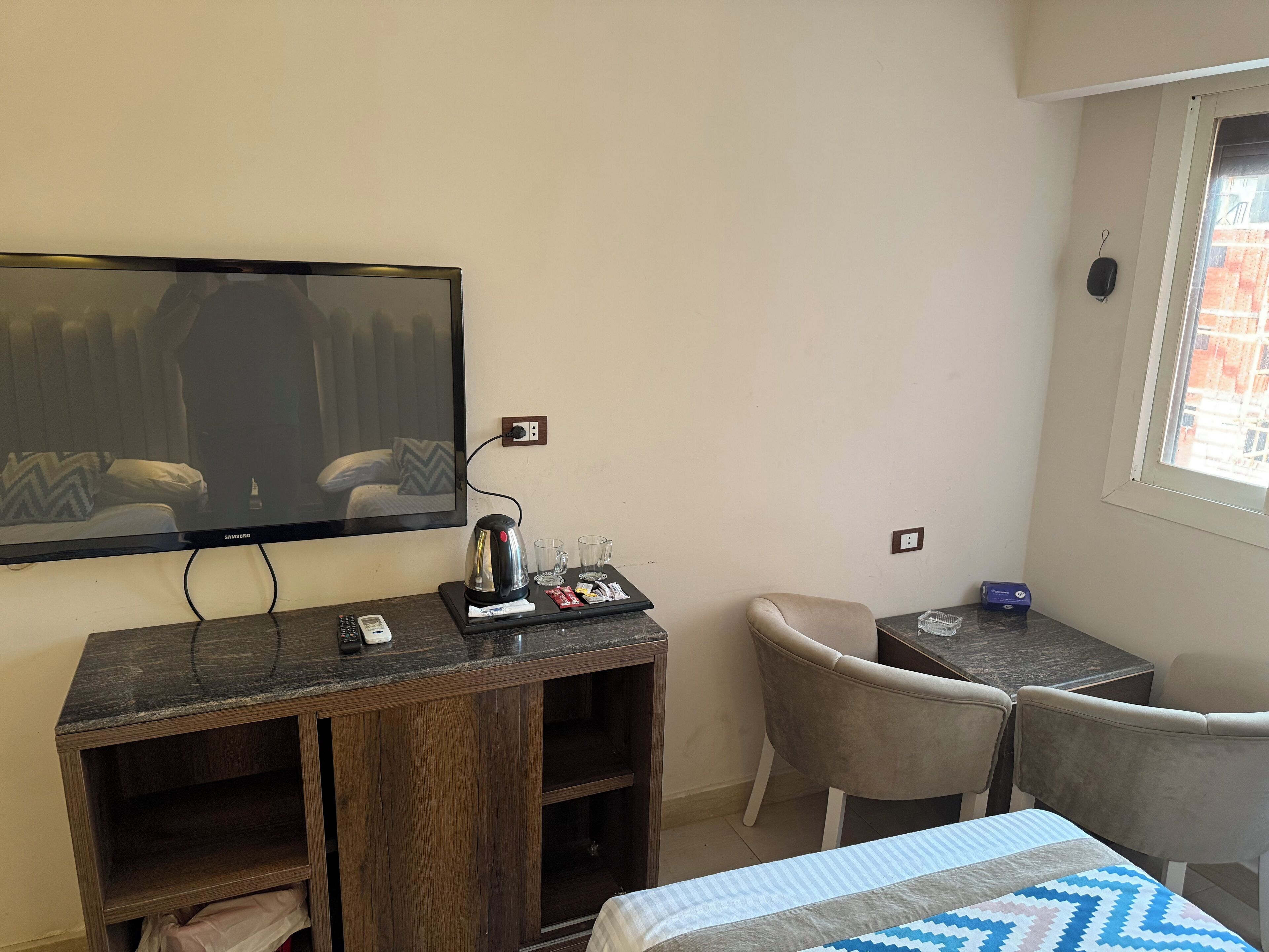 A hotel room with a bed, a TV mounted on the wall, a desk with a kettle, glasses, and a phone. There is a chair and a small table with a power outlet.