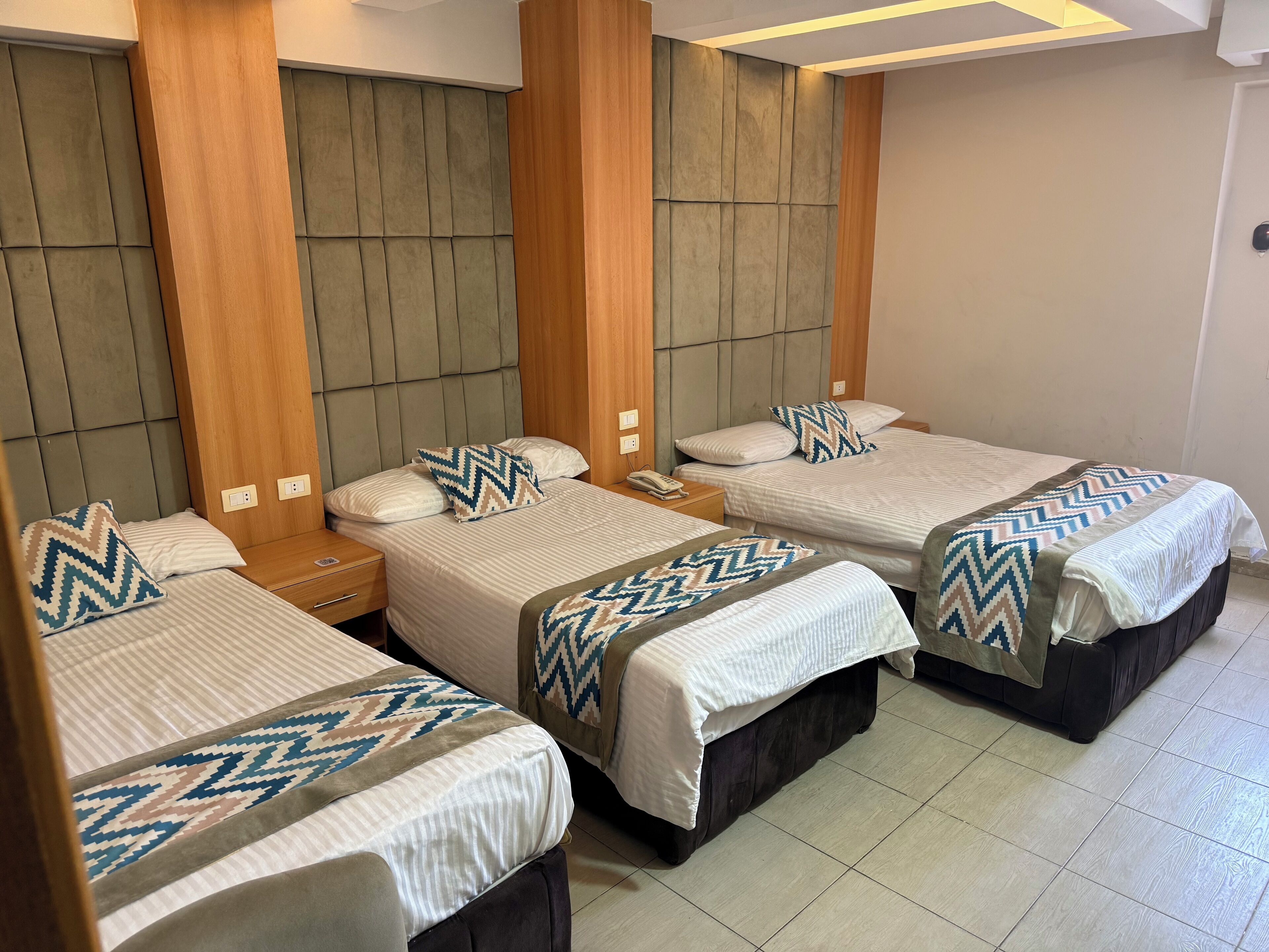 Quarto de hotel com quatro camas dispostas em linha, cada uma com colcha estampada e almofadas a combinar. O quarto possui cabeceiras de madeira e piso de azulejo.