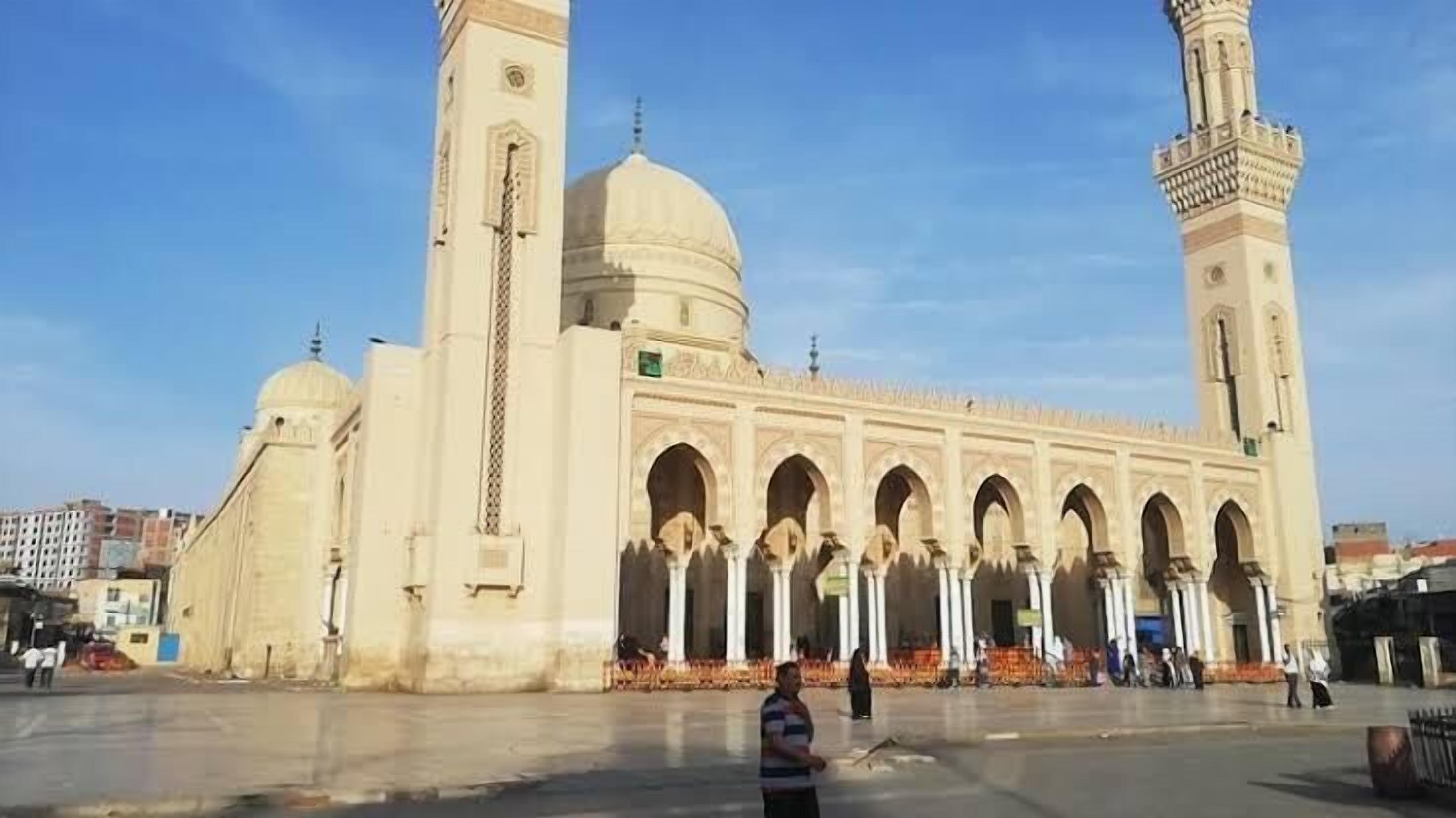 Una gran mezquita con cúpulas y minaretes, que cuenta con un patio con gente y algunos coches aparcados.