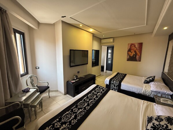 A hotel room with two beds, a desk, a chair, a television, and an air conditioning unit.