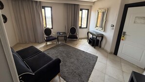Una sala de estar moderna con un sofá, dos sillones, una mesita y un escritorio. Hay dos ventanas con cortinas, una puerta y un espejo en la pared.