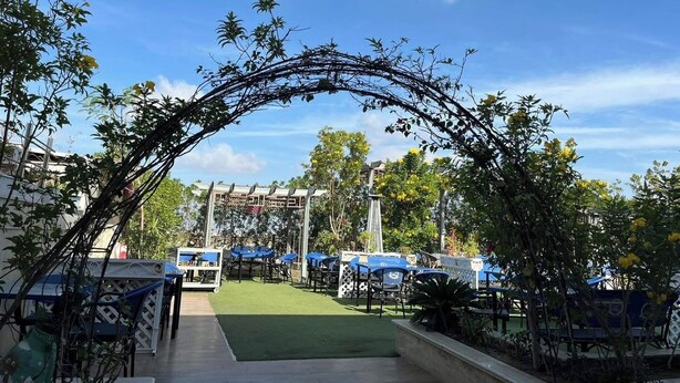 Un área de estar al aire libre con mesas y sillas, cubierta por un toldo azul, y un arco con plantas trepadoras.