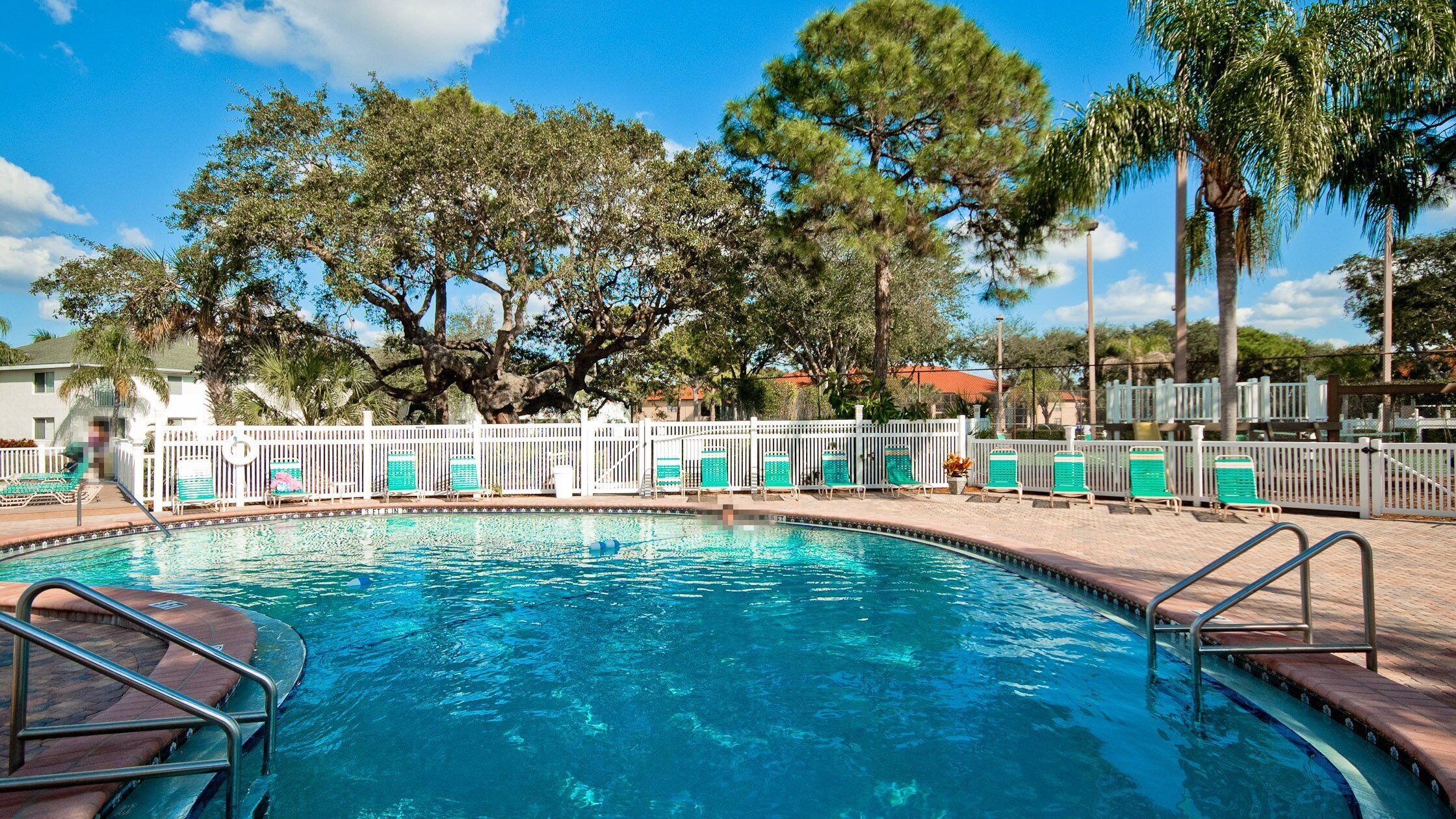 A swimming pool with a white fence, green lounge chairs, and trees in the background.