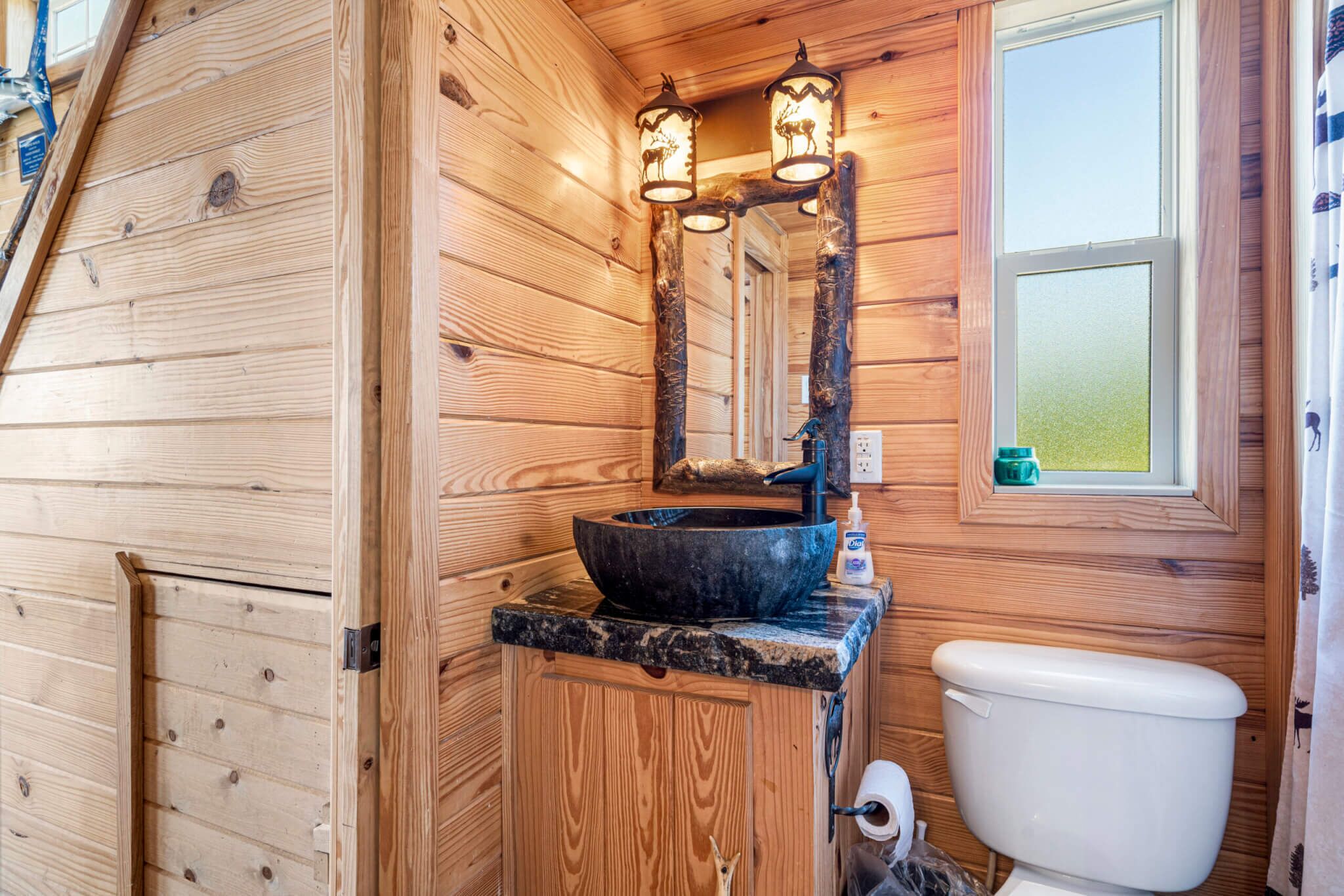 Baño de cabaña de madera con lavamanos de piedra, dos luces empotradas en la pared y una ventana.
