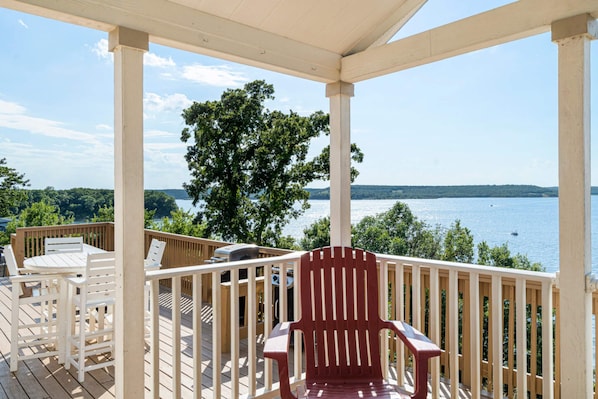 A wooden deck with a red chair and white table, overlooking a body of water and trees.