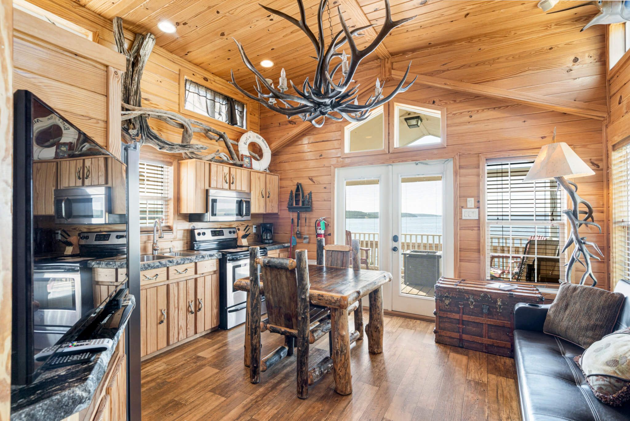 A log cabin interior with a wooden dining table, a sofa, and a large antler chandelier.