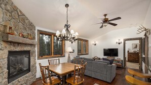 A spacious living area with a stone fireplace, a dining table with chairs, a grey sofa, a television, and a ceiling fan. - Cozy ski-in/ski-out home w/fireplace & clawfoot tub - Little Plume Flat (Big Sky)