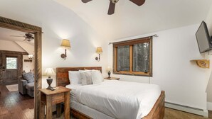 A bedroom with a wooden bed, two bedside tables with lamps, a ceiling fan, a window with a view, and a television on the wall. - Cozy ski-in/ski-out home w/fireplace & clawfoot tub - Little Plume Flat (Big Sky)
