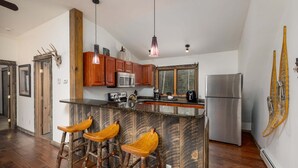 A modern kitchen with wooden cabinets, a stainless steel refrigerator, a bar area with stools, and a large window offering a view of the outdoors. - Cozy ski-in/ski-out home w/fireplace & clawfoot tub - Little Plume Flat (Big Sky)