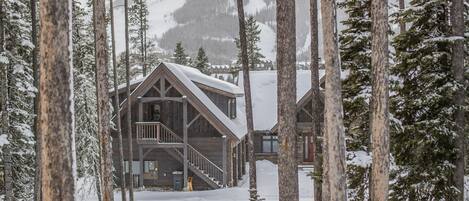 A wooden lodge with a balcony, surrounded by tall pine trees and a snowy mountain in the background.