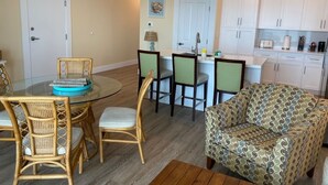 Un comedor con una mesa de vidrio y sillas de mimbre, un sofá estampado, y una cocina con gabinetes blancos y una lámpara colgante.