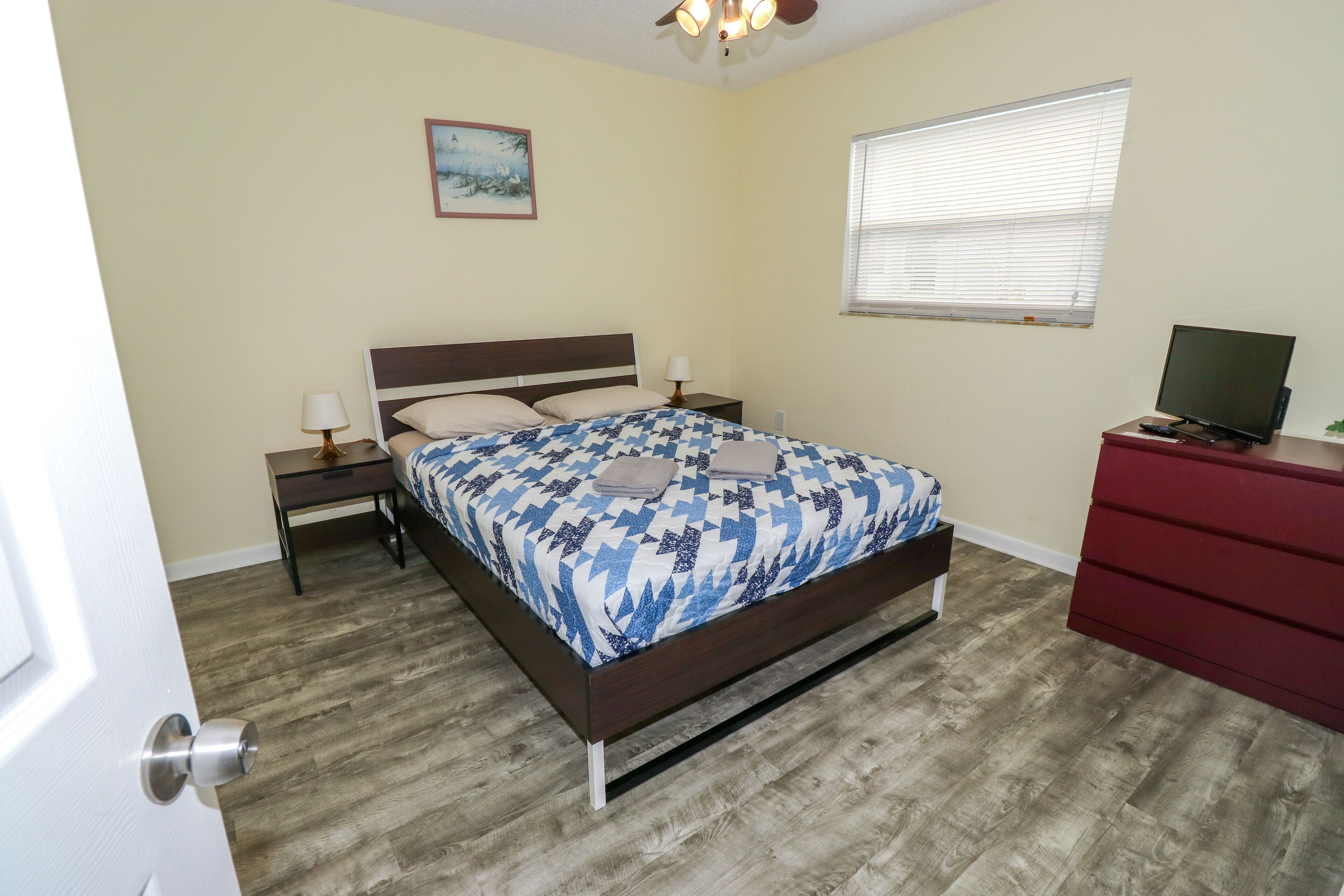 A bedroom with a bed, bedside tables, a TV, a window, and a painting on the wall.