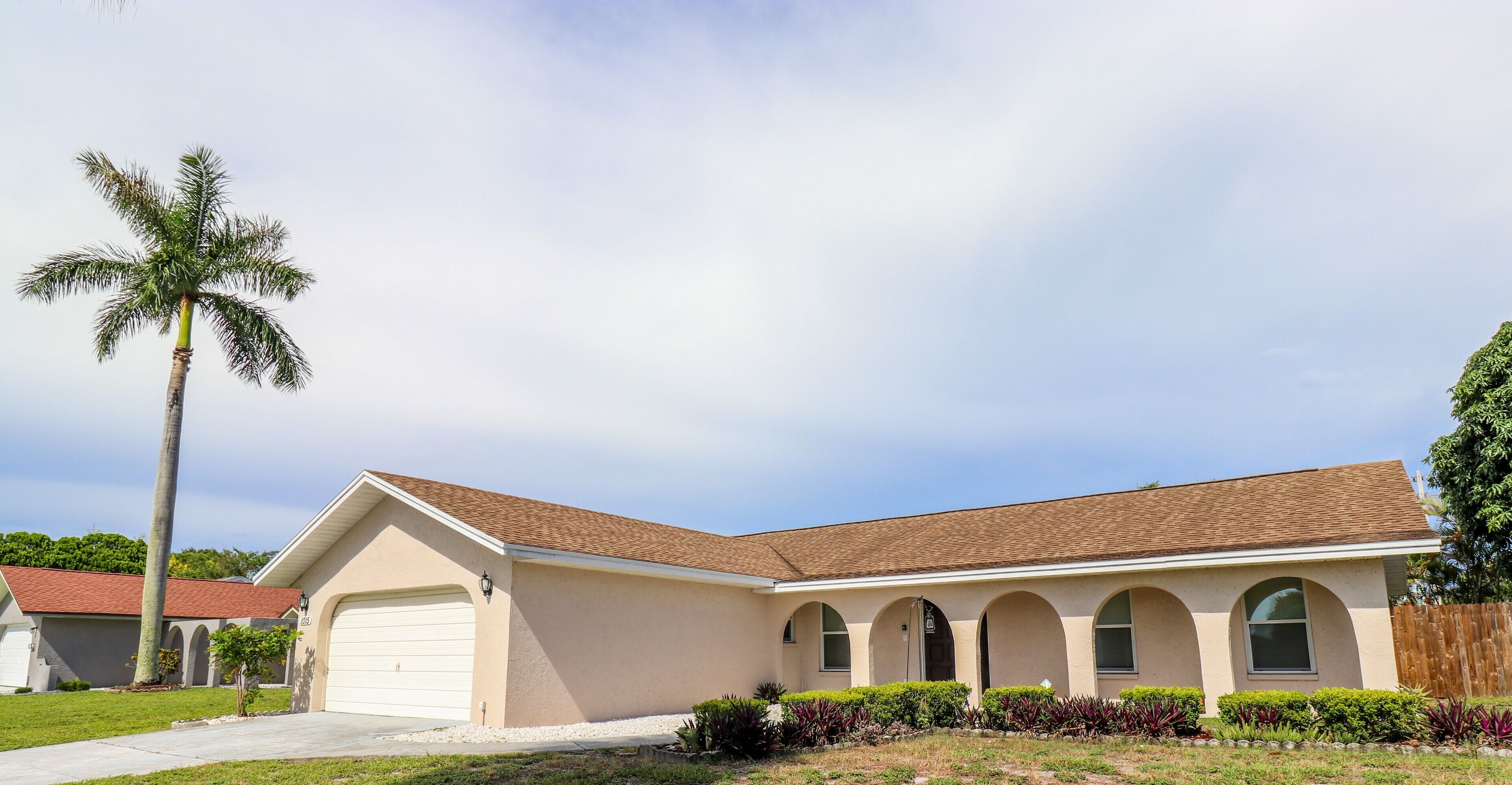 A single-story house with a beige exterior, a brown roof, and a garage. There is a palm tree to the left and a row of shrubs in front.